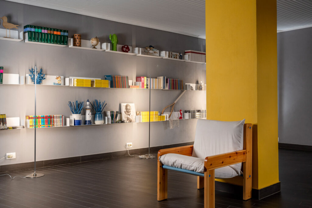 Salotto moderno con parete grigia, scaffali pieni di libri e decorazioni, e una sedia davanti a un pilastro giallo.