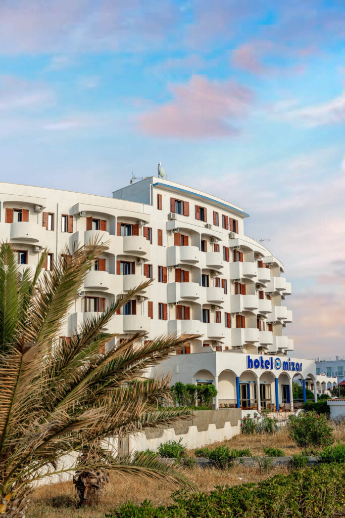 Edificio di un hotel bianco a più piani con balconi, circondato da vegetazione e cielo azzurro.