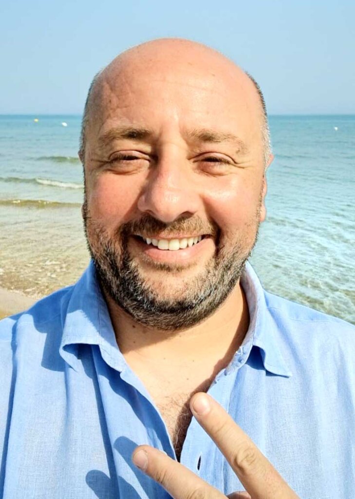 Uomo sorridente in spiaggia con camicia azzurra e gesto di pace.