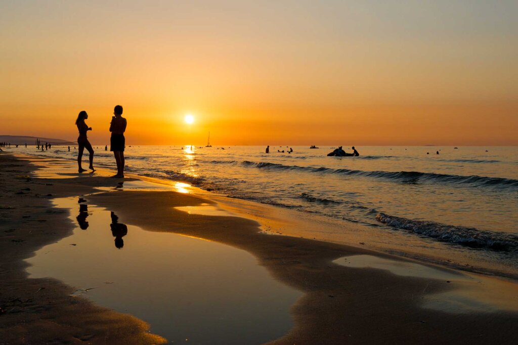 Tramonto sulla spiaggia con due persone in silhouette e riflessi sull'acqua.