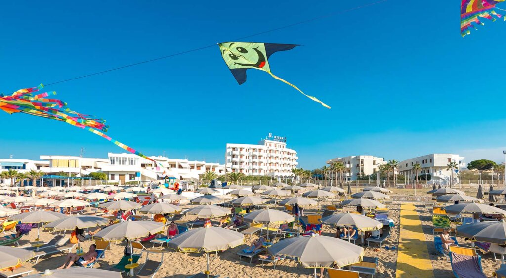 Spiaggia affollata con ombrelloni e un aquilone colorato nel cielo azzurro.